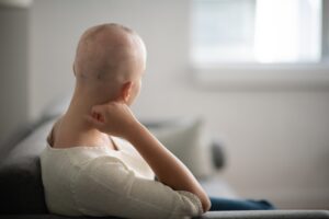 A woman is indoors in her living room. Her head is shaved due to chemotherapy. She is sitting and looking thoughtful.